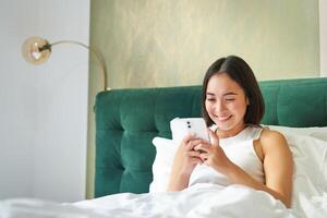 hermosa sonriente asiático mujer en cama, leyendo mensaje en teléfono inteligente y reír, disfrutando perezoso día en dormitorio, utilizando móvil teléfono foto