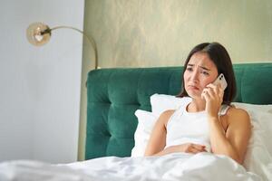 Portrait of concerned asian woman holding mobile phone, receive bad phone call, looking worried and upset, having difficult telephone conversation while lying in bed photo