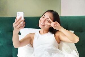 Positive girl blogger takes selfie in her bed, bedroom photo on smartphone, smiling and showing peace v-sign gesture