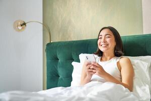 Beautiful smiling asian woman in bed, reading message on smartphone and laughing, enjoying lazy day in bedroom, using mobile phone photo