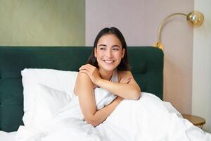 Happy mornings. Smiling asian woman wakes up in her bed, looks outside and feeld enthusiastic about her morning. Bedroom interior concept photo