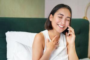 Close up portrait of cute asian girl in bed, talking on mobile phone with happy smiling face. Woman waking up and making a telephone call photo