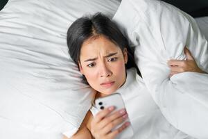 Portrait of asian girl wakes up in morning, looks at mobile phone with shocked face, overslept, hugging pillow and grimacing photo