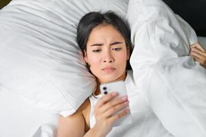 Close up portrait of asian girl lying in bed, looking at smartphone concerned, waking up late and staring at her alarm clock on mobile phone photo