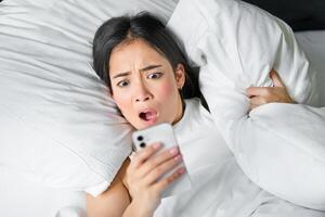 Close up portrait of asian girl lying in bed, looking at smartphone concerned, waking up late and staring at her alarm clock on mobile phone photo