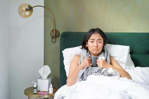 enfermo triste asiático niña acostado en su cama con gripe, captura un frío, tomando pastillas y Bebiendo caliente té, sensación trastornado foto