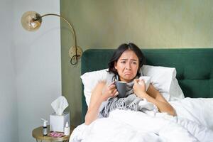 enfermo triste asiático niña acostado en su cama con gripe, captura un frío, tomando pastillas y Bebiendo caliente té, sensación trastornado foto