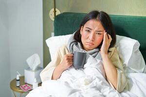retrato de asiático mujer con dolor de cabeza, atrapando columna, quedarse en enfermo salir a hogar, acostado en cama, Bebiendo caliente té, teniendo gripe foto