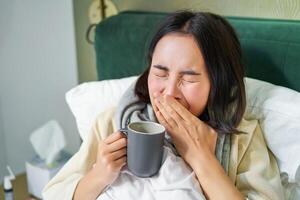 Close up of asian girl in bed, feeling sick, catching cold and sneezing, wiping runny nose, drinking hot tea photo