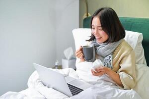 Portrait of asian sick girl, lying in bed with cold, having flu, talking to laptop, video chat, working from home on remote while on sick leave photo