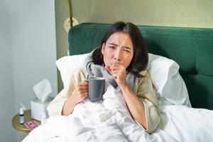 Portrait of couching asian girl in bed, feeling sick, catching cold and staying at home, looking unhealthy, drinking hot tea photo