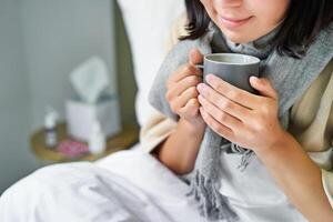 Close up of female hands holding hot drink, lying in bed, girl catching a cold and staying at home photo