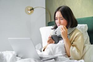 Portrait of asian girl sneezing and wiping runny nose, feeling sick, staying in bed with influenza, looking at laptop photo