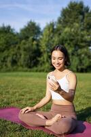 Portrait of asian girl sitting on mat in park, talking to smartphone, meditating online with yoga instructor on fresh air photo