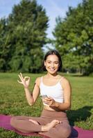 vertical Disparo de asiático mujer muestra bueno firmar, recomendando yoga formación en línea, meditación aplicación, haciendo ejercicios en Fresco aire en parque foto