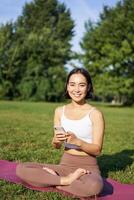 retrato de coreano niña meditando en parque, seguir yoga vídeo lección en teléfono inteligente aplicación, sentado en Fresco aire y relajante foto