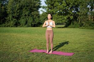 atención plena y bienestar. joven mujer haciendo yoga, en pie en estera en parque, haciendo asanas, meditando en Fresco aire, urbano vida concepto foto