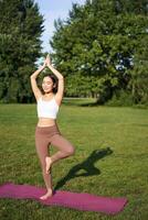 Vertical shot of asian woman standing in asana, doing yoga exercises on fresh air in park, wearing leggings, standing on rubber mat photo