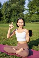 Vertical shot of asian woman shows okay sign and smartphone screen, recommending yoga training online, meditation app, doing exercises on fresh air in park photo