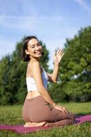 Portrait of asian girl on fitness class in park, sitting on rubber mat and wave hand at camera, say hello, meditating and practice yoga photo