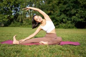 Young woman does yoga on lawn in park, stretching on fitness mat, wellbeing concept photo