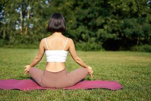Rear view of woman silhouette doing yoga, sitting on fitness mat and meditating on green lawn photo