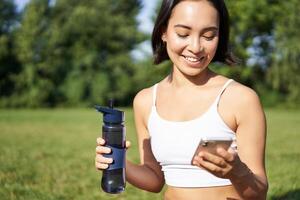 Smiling fitness girl drinks water, checks her app on smartphone and looks happy, stays hydrated on fresh air, sunny day in park photo