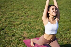 retrato de asiático joven mujer sonriente complacido, extensión su cuerpo en yoga asanas, en pie en estera y rutina de ejercicio en parque foto