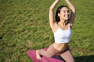 Portrait of asian young woman smiling pleased, stretching her body in yoga asana, standing on mat and workout in park photo