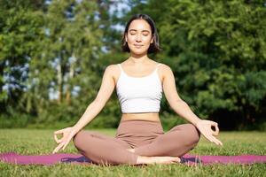 Sporty woman meditating on fresh air, sitting on fitness mat and practice yoga, smiling pleased. Sport and people concept photo