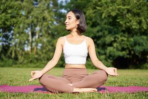 Sporty woman meditating on fresh air, sitting on fitness mat and practice yoga, smiling pleased. Sport and people concept photo