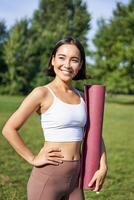 Vertical portrait of young asian fitness girl walks with rubber mat for yoga, goes on training session on fresh air in park, wears sports clothing photo