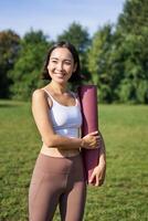 Vertical portrait of young asian fitness girl walks with rubber mat for yoga, goes on training session on fresh air in park, wears sports clothing photo