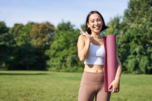 Happy asian fitness girl says hi, waves hand, stands with rubber yoga mat for workout in park, wears sport uniform photo