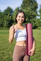 Excited young woman standing with sports mat, yoga clothes, shows okay sign, workout in park, wellbeing training session outdoors photo