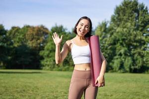 retrato de joven asiático mujer rutina de ejercicio en parque al aire libre, aptitud instructor olas mano y sonrisas, invita personas para yoga formación en Fresco aire foto