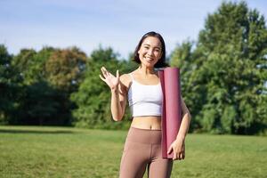 retrato de joven asiático mujer rutina de ejercicio en parque al aire libre, aptitud instructor olas mano y sonrisas, invita personas para yoga formación en Fresco aire foto