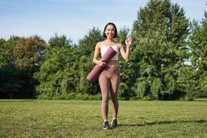 elegante deportista con caucho estera para yoga, dice Hola, olas a tú, Listo para rutina de ejercicio formación al aire libre en parque en césped foto