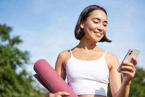 Portrait of smiling asian woman with yoga mat, looking at her smartphone and reading on application, standing in park wearing sport uniform photo