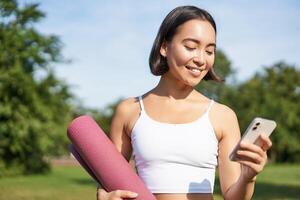 Portrait of smiling asian woman with yoga mat, looking at her smartphone and reading on application, standing in park wearing sport uniform photo