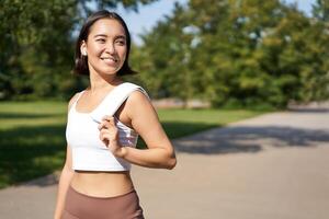 Smiling asian fitness girl holding towel on shoulder, workout in park, sweating after training exercises outdoors photo