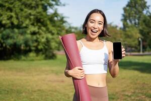 Smiling asian fitness girl with rubber yoga mat, shows her smartphone screen, recommends workout application, stands on lawn in park photo