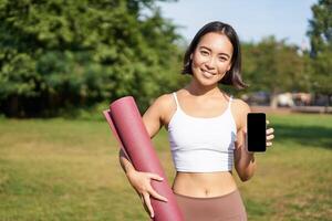 Smiling asian fitness girl with rubber yoga mat, shows her smartphone screen, recommends workout application, stands on lawn in park photo
