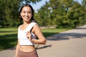 Smiling asian fitness girl holding towel on shoulder, workout in park, sweating after training exercises outdoors photo