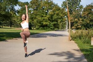 Excited young asian woman winning, finish running in park, saying yes, lifting hand up in triumph, celebrating victory or success photo