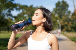 Happy asian sportswoman, runner drinks water from bottle while running, workout on fresh air in park photo