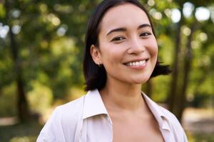 People concept. Smiling asian girl looking at camera with happy, joyful emotion, posing in green park on a warm summer day photo