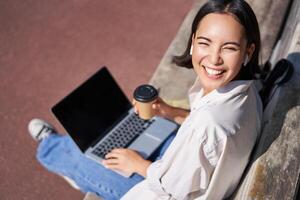 Beautiful young asian female student, sitting with laptop on bench in park, drinking takeaway coffee and listening music, working remotely, studying outdoors photo