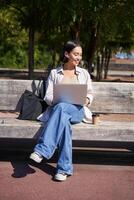 Portrait of korean woman sitting with laptop in park on bench, working outdoors, student doing homework, drinking takeaway coffee photo