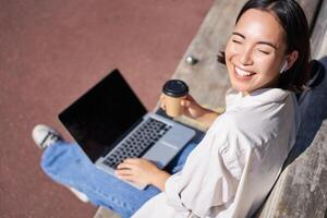 Beautiful young asian female student, sitting with laptop on bench in park, drinking takeaway coffee and listening music, working remotely, studying outdoors photo